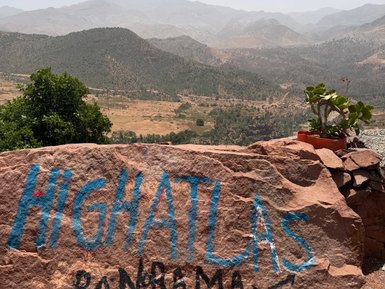 Ein großer Felsblock mit blauer und schwarzer Schrift zeigt den Namen "High Atlas Panorama" vor einer weiten Berglandschaft.