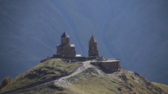 Auf einem grünen Hügel stehen zwei alte Kirchen, umgeben von majestätischen Bergen und einem klaren blauen Himmel.