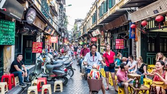 Eine belebte Gasse in Hanoi, gesäumt von Motorrädern und bunten Stühlen, mit Menschen, die sich unterhalten und essen.