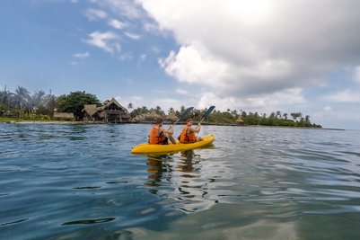 Zwei Personen in orangefarbenen Schwimmwesten paddeln in einem gelben Kajak auf ruhigem Wasser vor einer tropischen Kulisse.
