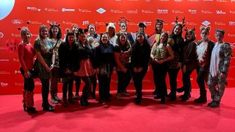 Eine Gruppe von Menschen in bunten Kostümen posiert fröhlich vor einer roten Wand mit Sponsorenlogos.