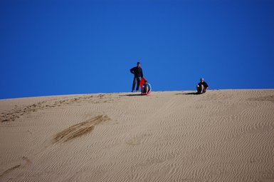 Drei Personen stehen und sitzen auf einer großen Sanddüne unter einem strahlend blauen Himmel, bereit für ein Abenteuer.