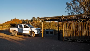 Ein weißes Fahrzeug steht an einem abgelegenen Campingplatz in Namibia, umgeben von sanften Hügeln und Bäumen.
