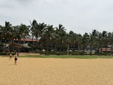 Zwei Kinder laufen am Strand entlang, umgeben von Palmen und einem Hotel im Hintergrund, unter einem bewölkten Himmel.