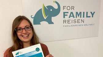 Eine lächelnde Frau hält einen Reiseflyer in der Hand, während sie vor einem Wandbild mit einem Elefanten steht.