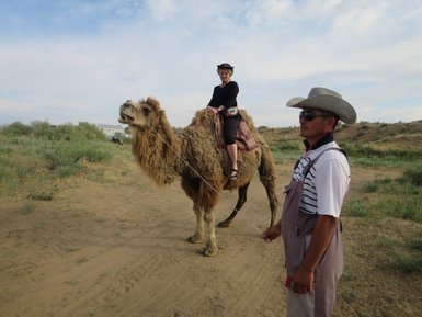 Eine Frau sitzt auf einem Kamel, während ein Mann mit einem Hut das Tier an der Leine hält, umgeben von einer grünen Landschaft.