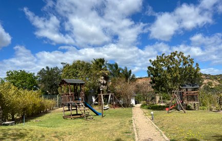 Ein Spielplatz im Freien mit Holzspielgeräten, umgeben von üppigem Grün und einem klaren blauen Himmel.