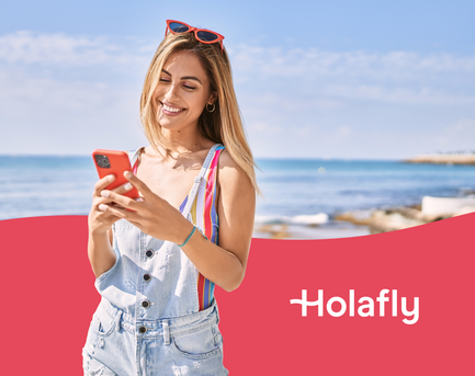 Eine junge Frau mit langen, blondierten Haaren steht am Strand und lächelt, während sie auf ihr rotes Handy schaut.