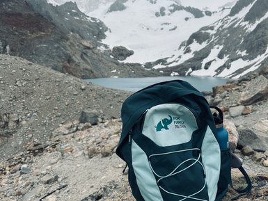 Ein Rucksack steht auf einem großen Stein, umgeben von schneebedeckten Bergen und einem klaren See im Hintergrund.