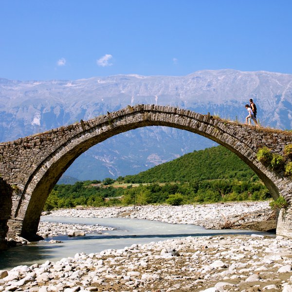 Ein Paar geht über eine alte Steinbrücke, umgeben von grünen Hügeln und majestätischen Bergen unter einem strahlend blauen Himmel.