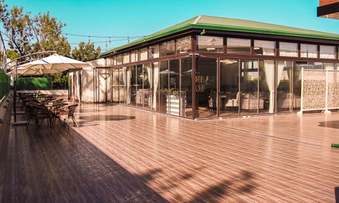 Eine sonnige Terrasse mit modernen Stühlen und Tischen, umgeben von grünen Pflanzen und einem klaren blauen Himmel.