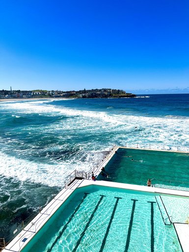 Der Pool am Bondi Beach reflektiert das Sonnenlicht, während Wellen an den Rand schlagen und Menschen schwimmen.