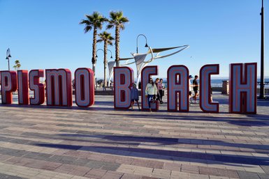 Große, bunte Buchstaben mit der Aufschrift 'Pismo Beach' stehen auf einer Promenade, umgeben von Palmen und Besuchern.