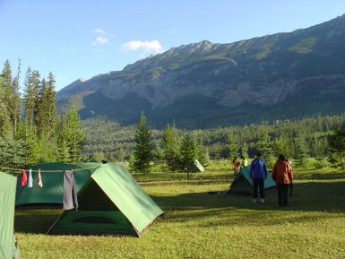 Zwei Personen stehen auf einem Campingplatz mit grünen Zelten, umgeben von hohen Bäumen und majestätischen Bergen im Hintergrund.