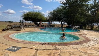 Ein Pool in der Natur mit mehreren Personen, die sich entspannen und schwimmen, umgeben von Bäumen und einem weiten Feld.