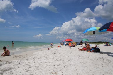 Ein sonniger Strand mit feinem, weißem Sand, wo Familien unter bunten Sonnenschirmen entspannen und spielen.