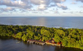 Das atemberaubende Bild zeigt eine friedliche Lodge am Wasser, umgeben von üppigem Grün und dem weiten Ozean im Hintergrund.