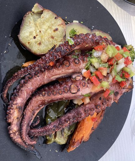 Gegrillter Oktopus liegt auf einem schwarzen Teller, garniert mit frischem Gemüse und Kräutern, umgeben von Süßkartoffeln.