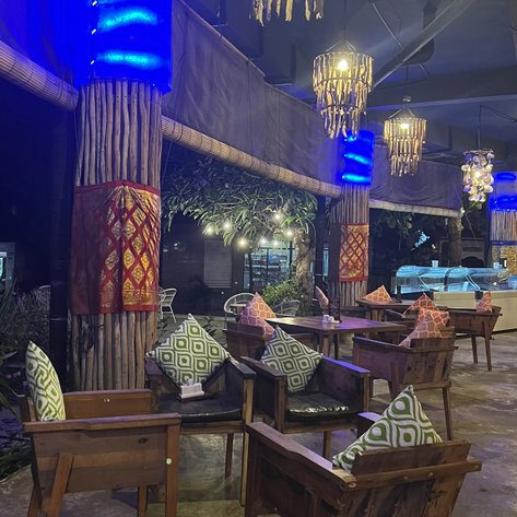 Ein stilvolles Restaurant mit Holzstühlen und bunten Kissen, beleuchtet von warmen und blauen Lichtern, schafft eine einladende Atmosphäre.