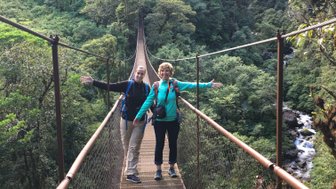 Zwei Wanderer stehen auf einer Hängebrücke, umgeben von üppigem Grün und einem Wasserfall im Hintergrund.