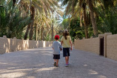 Zwei Kinder gehen Hand in Hand auf einem gepflasterten Weg, umgeben von hohen Palmen und einem sonnigen Himmel.