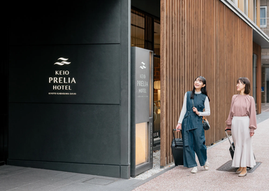 Zwei Frauen stehen vor dem Keio Prelia Hotel, lächelnd und mit Gepäck, bereit für ihren Aufenthalt in Kyoto.