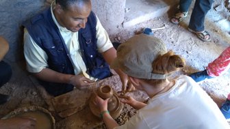 Ein Mann und eine Frau arbeiten gemeinsam an einem Tontopf in einer traditionellen Töpferei, umgeben von Erde und Werkzeugen.
