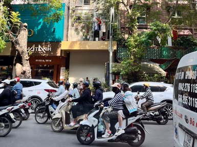 Eine belebte Straße in Hanoi, auf der viele Motorräder und Autos im Verkehr stehen, umgeben von grünen Bäumen.