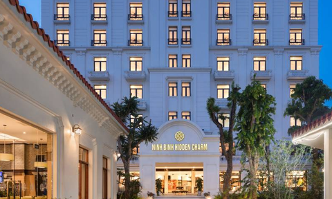Ein elegantes Hotelgebäude mit dem Namen 'Ninh Binh Hidden Charm' strahlt in der Dämmerung, umgeben von üppigem Grün.