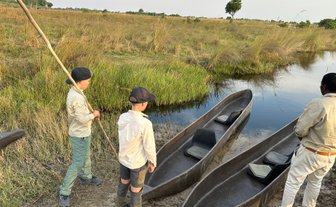 Drei Kinder und ein Erwachsener stehen am Ufer eines ruhigen Gewässers, umgeben von hohem Gras und Booten.