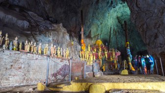 In einer Höhle stehen zahlreiche goldene Buddha-Statuen, umgeben von bunten Tüchern und einer mystischen Atmosphäre.