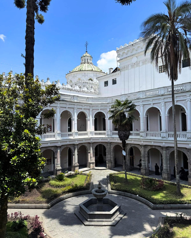 Ein eleganter Innenhof mit einem zentralen Brunnen, umgeben von weißen Säulen und üppigem Grün unter einem strahlend blauen Himmel.