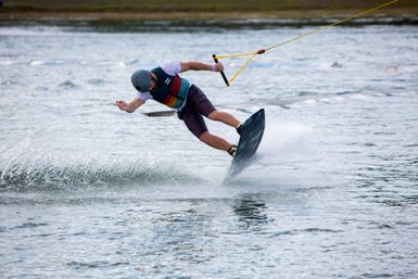 Ein Wakeboarder springt über das Wasser, während er mit einem Seil von einem Boot gezogen wird, umgeben von spritzendem Wasser.