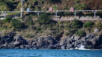 Eine farbenfrohe Seilbahn fährt über eine grüne Küstenlandschaft, während Wellen sanft gegen die Felsen schlagen.