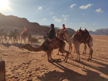 Eine Gruppe von Kamelen mit Reitern durchquert die Wüste, während die Sonne am Horizont strahlt und Schatten wirft.