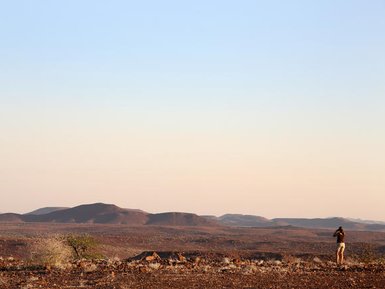 Ein Reisender steht auf einer kargen Landschaft und betrachtet die weite, hügelige Wüste im Damaraland bei Sonnenuntergang.