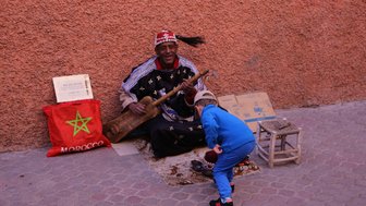 Ein lächelnder Musiker in traditioneller Kleidung spielt ein Instrument, während ein kleiner Junge neugierig daneben steht.