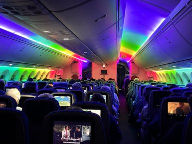 Der Innenraum eines Flugzeugs ist in bunten Lichtern beleuchtet, während Passagiere Filme auf Bildschirmen ansehen.
