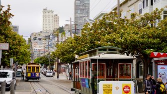 Eine Straßenbahn fährt durch eine belebte Straße in San Francisco, umgeben von hohen Gebäuden und üppigem Grün.