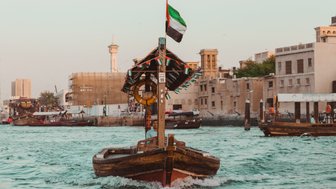 Ein traditionelles Holzboot fährt durch das Wasser des Dubai Creeks, umgeben von modernen und historischen Gebäuden.