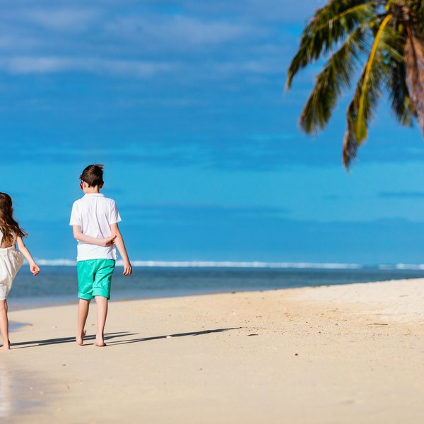 Eine Mutter und zwei Kinder spazieren barfuß am Strand entlang, umgeben von Palmen und einem strahlend blauen Himmel.