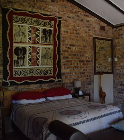Ein gemütliches Schlafzimmer mit einem großen Bett, bunten Kissen und einer kunstvollen Wanddekoration aus afrikanischen Motiven.