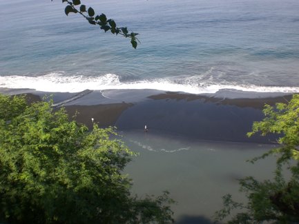 Ein Blick auf eine ruhige Lagune mit schwarzem Sandstrand, umgeben von üppigem Grün und sanften Wellen des Ozeans.