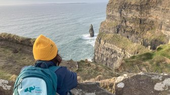 Eine Person mit einer gelben Mütze blickt auf die beeindruckenden Klippen von Moher und das rauschende Meer darunter.