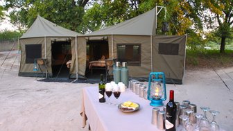 Zwei große Zelte stehen in einer sandigen Umgebung, umgeben von Bäumen, mit einem Tisch voller Getränke und Snacks.