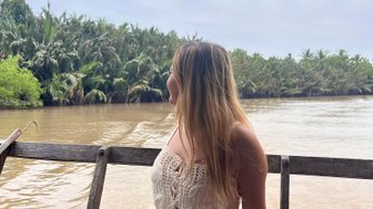 Eine Frau mit langen Haaren sitzt auf einem Holzboot und blickt auf den ruhigen Mekongfluss, umgeben von üppigem Grün.