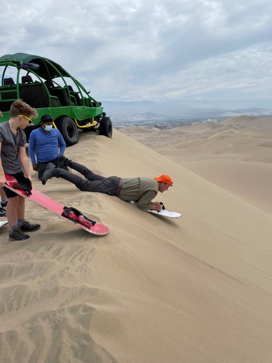 Eine Gruppe von Menschen steht an einer Sanddüne, während einer von ihnen auf einem Board liegt und sich auf die Abfahrt vorbereitet.