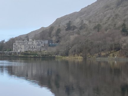 Ein majestätisches Schloss spiegelt sich im ruhigen Wasser eines Sees, umgeben von sanften Hügeln und kahlen Bäumen.