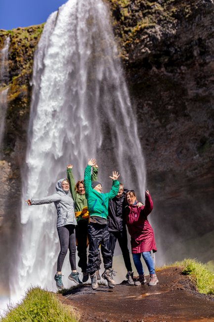 Sechs fröhliche Menschen springen vor dem beeindruckenden Seljalandsfoss-Wasserfall in Island in die Luft.