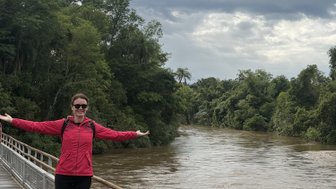 Eine Frau in einem pinken Kleid steht auf einer Holzplanke und breitet die Arme aus, um den malerischen Fluss zu genießen.